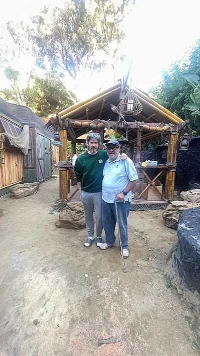 Two men standing in front of a completed bamboo garden structure with a shingled roof and hanging lanterns